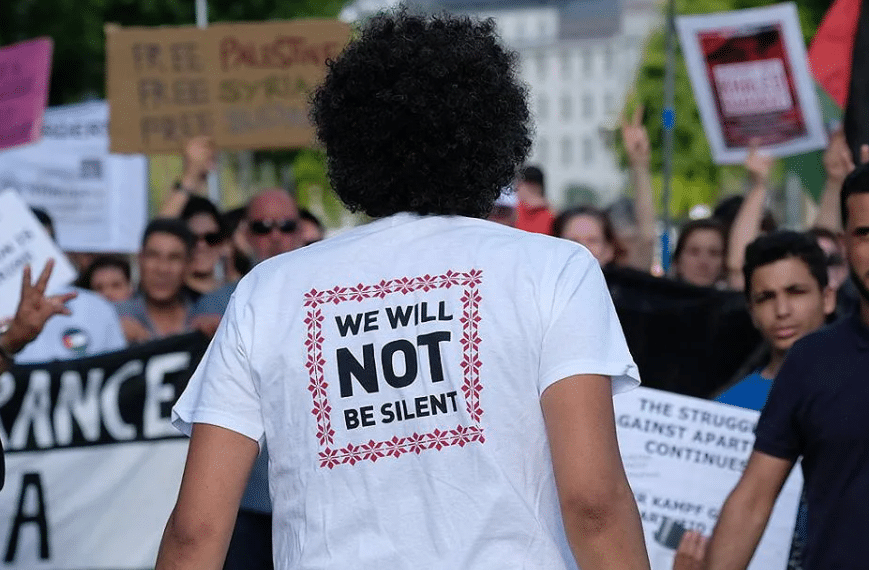 The back of a man's tshirt at a march, which reads 'We Will Not Be Silent'